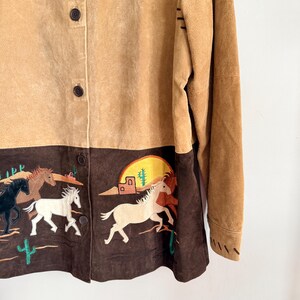 May include: Tan suede button-up jacket with a brown embroidered bottom panel. The panel features running horses, cacti, and a desert scene with a setting sun. The jacket has long sleeves and brown buttons.