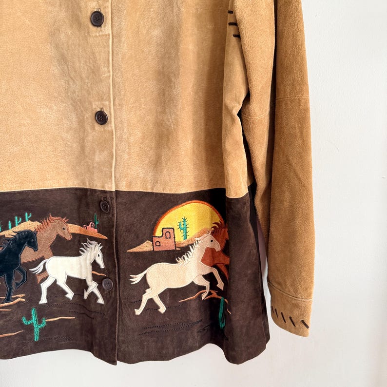 May include: Tan suede button-up jacket with a brown embroidered bottom panel. The panel features running horses, cacti, and a desert scene with a setting sun. The jacket has long sleeves and brown buttons.