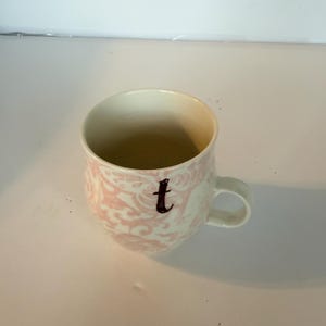 May include: A white ceramic mug with a pink floral pattern and a black letter "t". The mug has a rounded shape with a handle and a cream-colored interior. The design is a classic, elegant style.