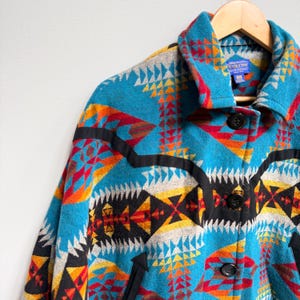May include: A Pendleton wool jacket featuring a vibrant geometric pattern in turquoise, red, orange, yellow, and black. The jacket has a pointed collar, three black buttons down the front, and black trim accents across the chest and pockets.
