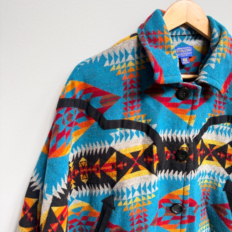 May include: A Pendleton wool jacket featuring a vibrant geometric pattern in turquoise, red, orange, yellow, and black. The jacket has a pointed collar, three black buttons down the front, and black trim accents across the chest and pockets.