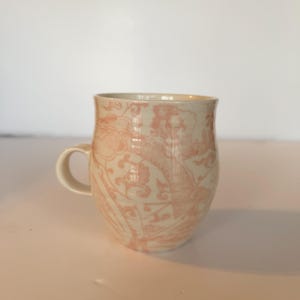 May include: A ceramic mug with a cream-colored base and a pink floral pattern. The mug has a rounded body, a curved handle, and a slightly flared rim. The floral design is a repeating pattern of stylized flowers and leaves.