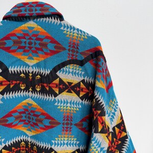 May include: A long-sleeved shirt featuring a vibrant geometric pattern in shades of turquoise, red, orange, yellow, black, and grey. The garment includes a pointed black yoke across the shoulders and a fold-over collar, showcasing a woven texture.