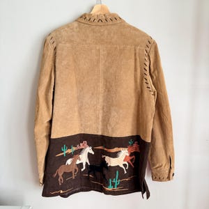May include: Tan suede jacket with embroidered horses and cacti on the lower back panel. The jacket has a button-down front, long sleeves, and decorative stitching along the seams and collar.