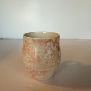 May include: A ceramic cup with a cream-colored base and a pink floral pattern. The cup has a rounded shape and a wide opening. The pink pattern features stylized floral and bird motifs. The cup is likely used for drinking beverages.