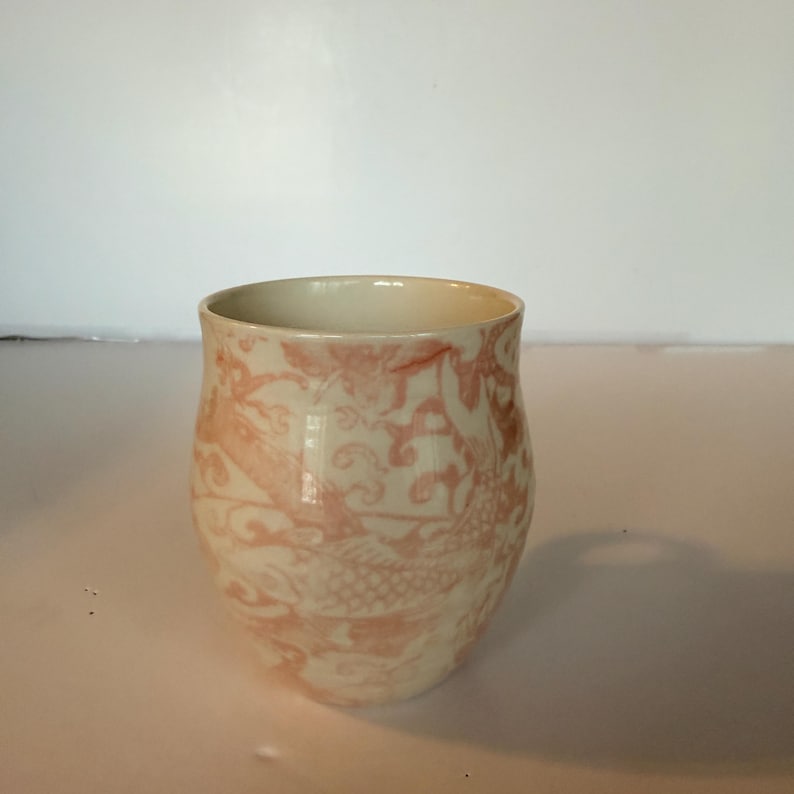 May include: A ceramic cup with a cream-colored base and a pink floral pattern. The cup has a rounded shape and a wide opening. The pink pattern features stylized floral and bird motifs. The cup is likely used for drinking beverages.