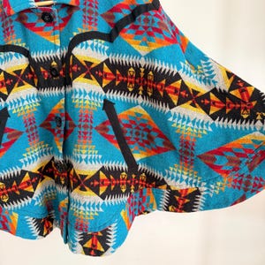 May include: A vibrant cape featuring a geometric pattern in shades of turquoise, red, yellow, black, and white. The garment includes a collared neckline, a front button closure with black buttons, and black trim accents along the chest and pockets.