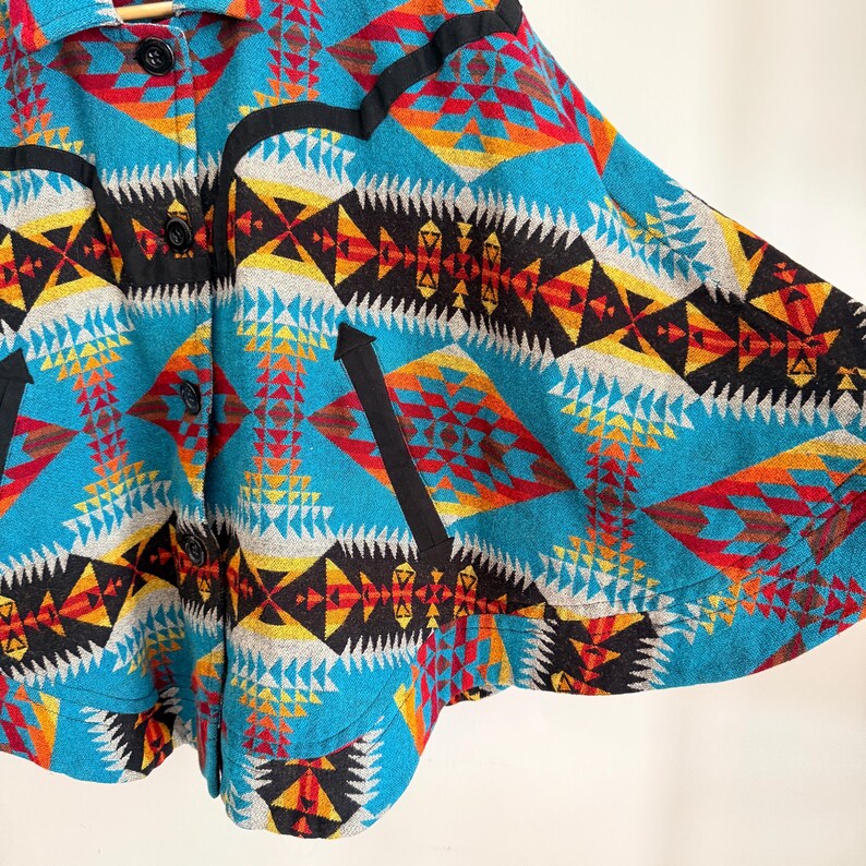 May include: A vibrant cape featuring a geometric pattern in shades of turquoise, red, yellow, black, and white. The garment includes a collared neckline, a front button closure with black buttons, and black trim accents along the chest and pockets.