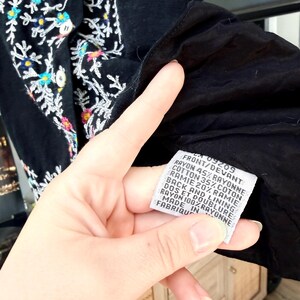 May include: Black garment with colorful floral embroidery. The label reads "FRONT/DEVANT RAYON 45% RAYONNE COTTON 35% COTON RAMIE 20% RAMIE BACK AND LINING RAYON 100% RAYONNE".