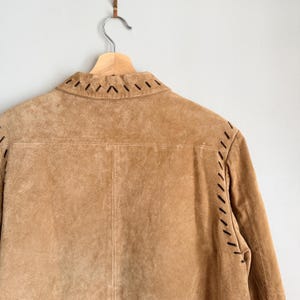 May include: A tan suede jacket hangs on a wooden hanger. The jacket features decorative stitching in black along the collar and seams. The back of the jacket is visible, showcasing the texture and color of the suede material.