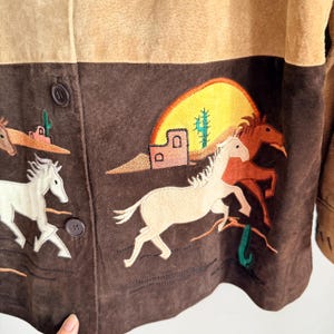 May include: A suede jacket with a two-tone design in tan and brown. The lower section features embroidered desert scenes with horses, cacti, and a setting sun. The jacket has button closures.