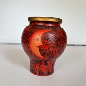 Könnte beinhalten: Kleine, rote Vase mit strukturierter Oberfläche und goldenem Rand. Die Vase zeigt einen bemalten Halbmond mit einem Gesicht in Orange- und Cremetönen. Die Vase hat eine einzigartige, abgerundete Form.