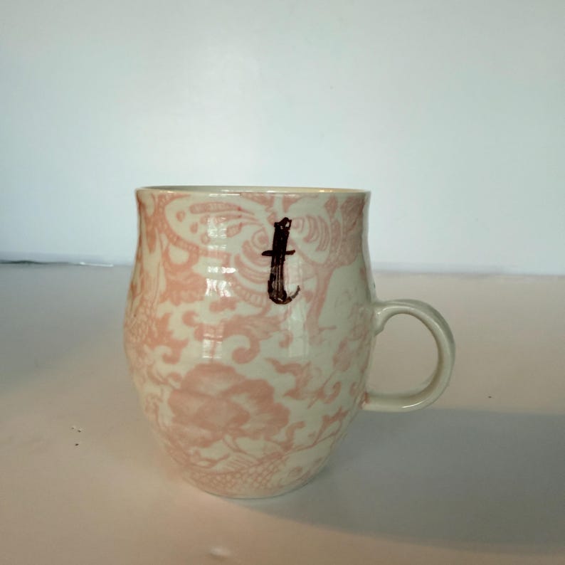 May include: A ceramic mug with a cream-colored base and a pink floral pattern. The mug has a curved shape, a handle, and a black letter "t" on the front. The mug is on a white surface.