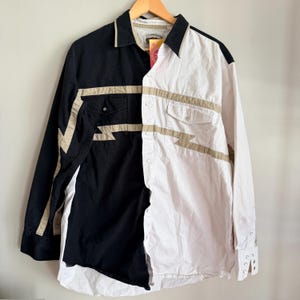 Puede incluir: Camisa abotonada de manga larga, dividida en negro y blanco. Acentos de zigzag y rayas beige adornan la camisa, con dos bolsillos en el pecho. La camisa está colgada en una percha de madera.