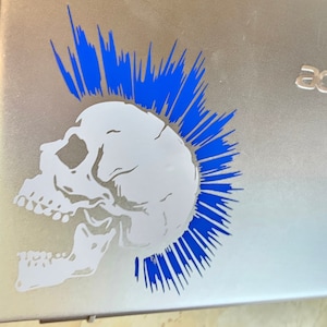 Peut inclure: Un crâne blanc avec une crête bleue orne un ordinateur portable argenté. Le crâne est un graphisme, et la crête est faite de lignes pointues. Le logo "acer" est visible en haut à droite de l'ordinateur.