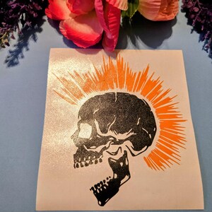 May include: A black skull with an orange mohawk design on a white background. The skull is facing left with its mouth open. The image is on a light blue surface with flowers in the background.