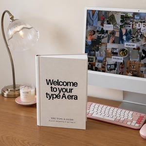 以下が含まれることがあります： ベージュのハードカバー本。表紙には黒い文字で「Welcome to your type A era」および「The Type A Guide Plan it. Prioritize it. Become it.」と記されており、木製のデスクの上でコンピューターモニター、ピンクのキーボード、マウス、ランプの隣に置かれています。