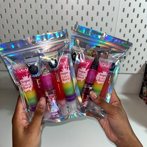 Puede incluir: Dos bolsas holográficas llenas de tubos de brillo labial. Cada bolsa contiene múltiples tubos con etiquetas coloridas, incluyendo el texto "Sugar Color Burst" y "HP Shine". Los tubos de brillo labial son de varios tonos de rosa, rojo y transparente.