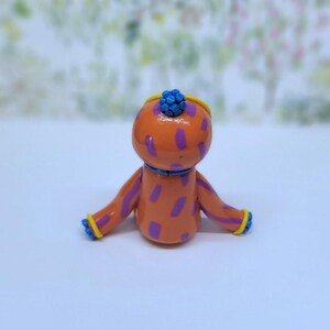 May include: A quirky orange figurine with purple spots, blue detailing, and a blue floral headpiece. The arms and head are trimmed with yellow. The figure is set against a soft, blurred backdrop.