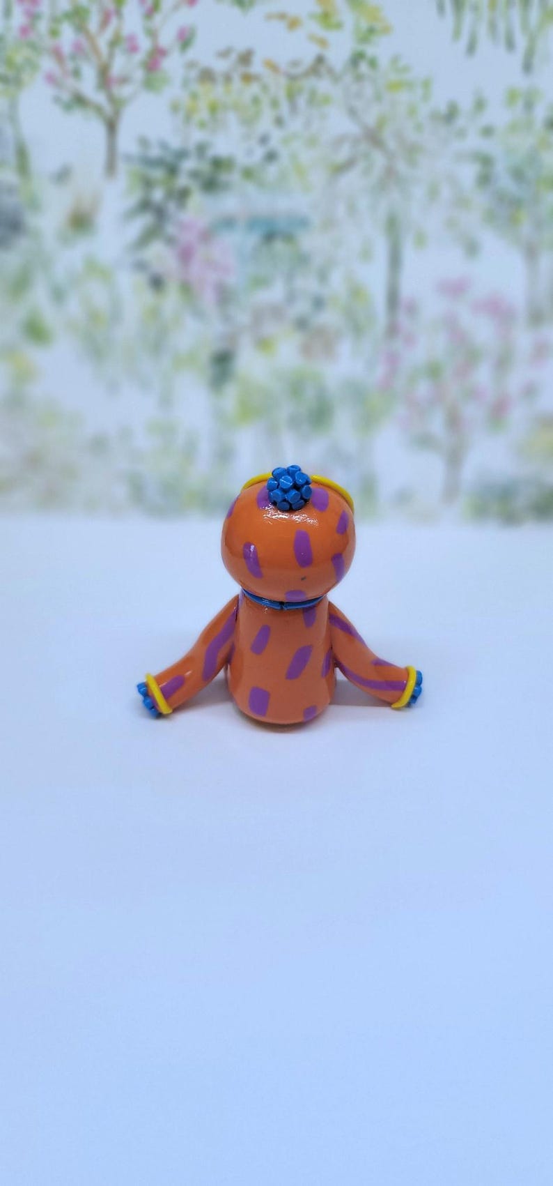 May include: A quirky orange figurine with purple spots, blue detailing, and a blue floral headpiece. The arms and head are trimmed with yellow. The figure is set against a soft, blurred backdrop.