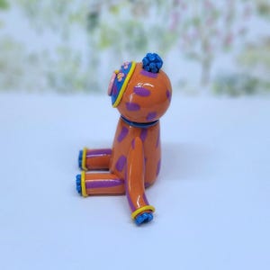 May include: A colourful orange dog figurine with purple spots, blue details, and a hat. The figure is seated, with a blurred backdrop of trees and flowers. The figurine is a decorative item.