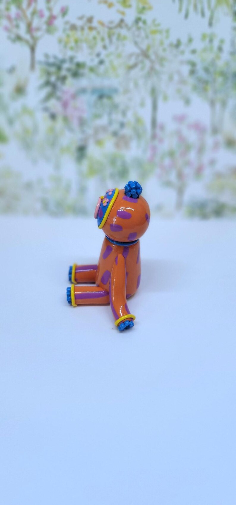 May include: A colourful orange dog figurine with purple spots, blue details, and a hat. The figure is seated, with a blurred backdrop of trees and flowers. The figurine is a decorative item.