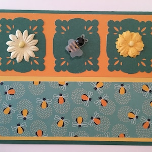 May include: A green card with a yellow, orange, and green border. The card has three cut-out squares with a white flower, a bee, and a yellow flower in each square. The bottom of the card has a green background with a pattern of white dots and bees.
