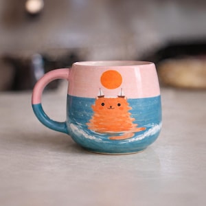 Puede incluir: Una taza de cerámica con un diseño caprichoso. La mitad superior es rosa claro, la mitad inferior es azul con detalles de olas blancas. Un gato naranja flota en el agua, con un sol encima. El asa es rosa y azul.