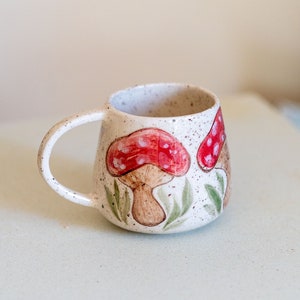 Pode incluir: Uma caneca de cerâmica salpicada com um design de cogumelos caprichoso. Os cogumelos têm chapéus vermelhos com manchas brancas e caules castanhos, rodeados por folhas verdes. A caneca tem uma alça curva e uma abertura larga.