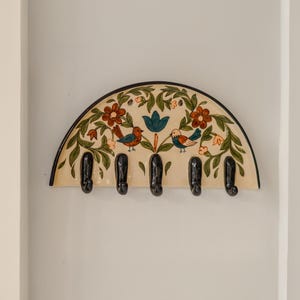 Op de afbeelding: Een decoratieve wandgarderobe met een halfrond ontwerp. De garderobe heeft een crèmekleurige achtergrond met een bloemen- en vogelmotief in rood, groen en blauw. Het heeft zes zwarte haken om items op te hangen.