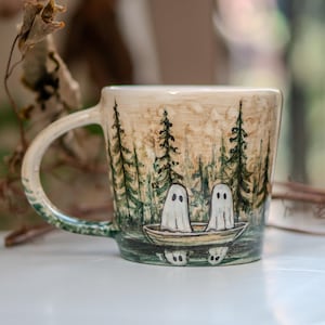 Puede incluir: Una taza de cerámica con una escena de bosque y dos figuras de fantasmas en un bote. La taza tiene un asa y está pintada con colores verdes, marrones y blancos. El diseño incluye árboles y una masa de agua.