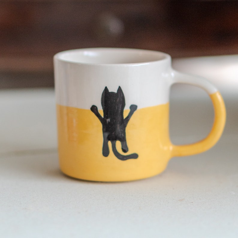 May include: A ceramic mug with a white top half and a yellow bottom half. A black cat illustration is centered on the yellow section. The handle and the rim of the mug are white.