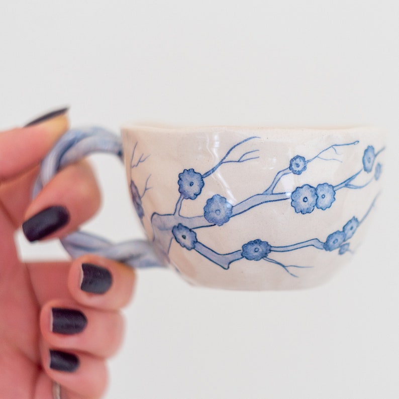 May include: A ceramic teacup with a twisted handle, featuring a hand-painted design of blue floral branches on a cream-colored background. The cup is held by a hand with dark nail polish, showcasing the delicate artistry of the teacup.