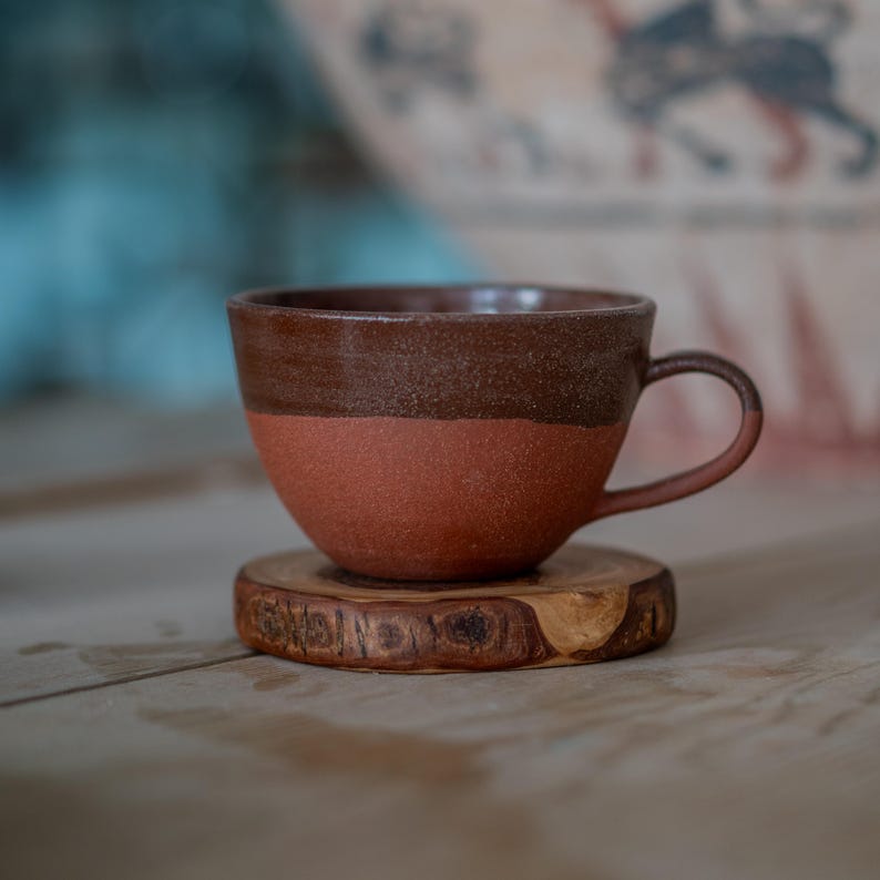 May include: A handcrafted ceramic mug, featuring a dark brown upper section and a reddish-brown lower section. It rests on a small, round wooden coaster. The mug has a curved handle and a rustic, textured finish.