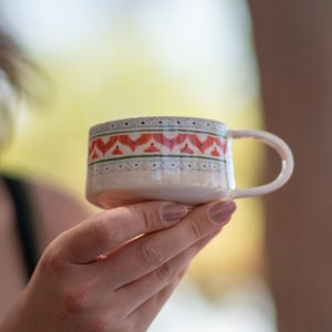 Könnte beinhalten: Eine kleine, weiße Keramiktasse mit einem dekorativen Band aus roten, grünen und blauen Mustern. Die Tasse hat einen abgerundeten Henkel und einen leicht ausgestellten Rand. Das Design ist handbemalt und verleiht ihm ein einzigartiges, handwerkliches Gefühl.