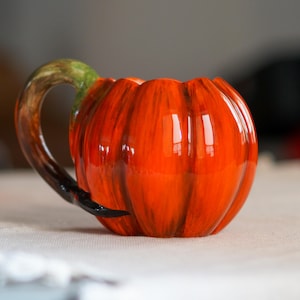 May include: A ceramic mug shaped like a pumpkin, with a glossy orange finish and vertical grooves. The handle is a curved stem with green and brown coloring. A black accent is at the base of the handle.