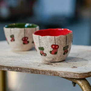 Pode incluir: Duas tigelas de cerâmica com exterior salpicado em branco sujo. O interior de uma tigela é verde, a outra vermelha. Cada tigela é decorada com desenhos de meias de Natal penduradas em vermelho e verde.