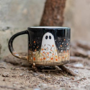 May include: Black ceramic mug featuring a white ghost with black eyes, surrounded by orange flowers and green grass. The mug has a black background and a curved handle. The mug is sitting on a natural surface.