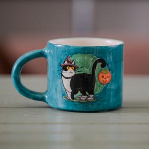 以下が含まれることがあります： 魔女の帽子をかぶり、カボチャのランタンを持った黒猫のイラストが描かれた、ターコイズ色のセラミックマグカップ。白い内側とターコイズ色のハンドルが付いています。ハロウィンに最適です。