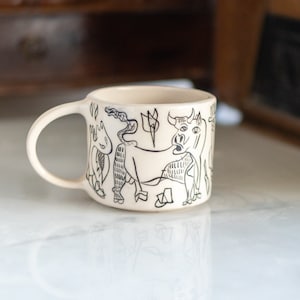May include: Cream-colored ceramic mug with a black line drawing of a bull and other abstract figures. The mug has a curved handle and a cylindrical shape. The design is reminiscent of folk art.