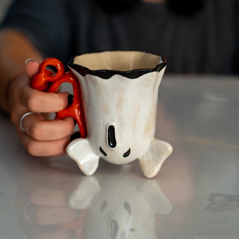 K&ouml;nnte beinhalten: Eine skurrile Keramik-Tasse mit wei&szlig;em Korpus, schwarzem Rand und einem leuchtend roten Henkel. Die Tasse hat ein einzigartiges Design mit kleinen, abgerundeten F&uuml;&szlig;en und einem schwarzen vertikalen Akzent. Die Tasse wird von einer Person gehalten.