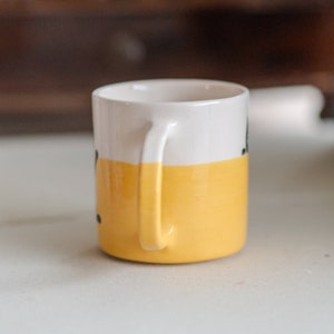 May include: A small ceramic mug with a white top half and a yellow bottom half. The mug has a curved handle and a simple design. The mug is sitting on a white surface.
