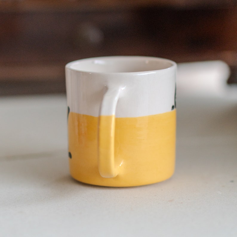 May include: A small ceramic mug with a white top half and a yellow bottom half. The mug has a curved handle and a simple design. The mug is sitting on a white surface.