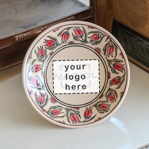Könnte beinhalten: Dekorative Keramikplatte mit cremefarbenem Grund und floralem Design. Der Rand zeigt rote Tulpen und grüne Blätter. Die Mitte hat einen weißen Bereich mit dem Text "your logo here".