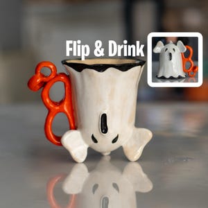 K&ouml;nnte beinhalten: Eine wei&szlig;e, keramikartige, geisterf&ouml;rmige Tasse mit einem orangefarbenen Griff. Die Tasse hat schwarze Akzente und ein l&auml;chelndes Gesicht. Die Worte "Flip & Drink" stehen &uuml;ber der Tasse.