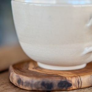 May include: A cream-coloured ceramic teacup with a rounded base sits on a small, round wooden coaster. The cup has a textured surface and a handle. The coaster is made of wood with a natural grain and a dark, irregular edge.