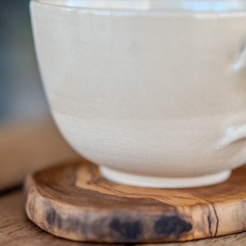 May include: A cream-coloured ceramic teacup with a rounded base sits on a small, round wooden coaster. The cup has a textured surface and a handle. The coaster is made of wood with a natural grain and a dark, irregular edge.