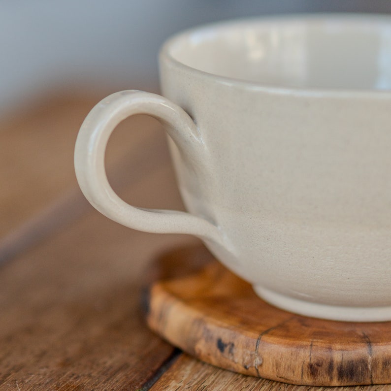 May include: A cream-coloured ceramic mug with a rounded handle. The mug sits on a small, round wooden coaster with a natural wood grain pattern. The mug's surface has a slightly textured appearance, and the background is blurred.