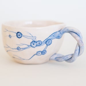 May include: A handmade ceramic teacup with a twisted handle. The cup is off-white with blue floral branch designs. The handle is a twisted rope design in blue and white. The cup is perfect for enjoying tea or coffee.