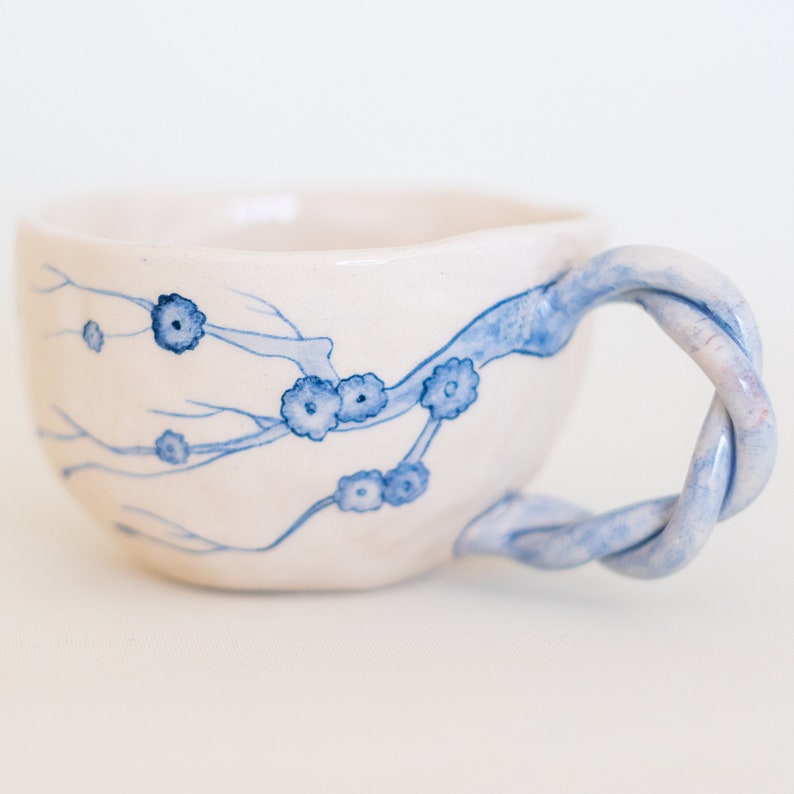 May include: A handmade ceramic teacup with a twisted handle. The cup is off-white with blue floral branch designs. The handle is a twisted rope design in blue and white. The cup is perfect for enjoying tea or coffee.